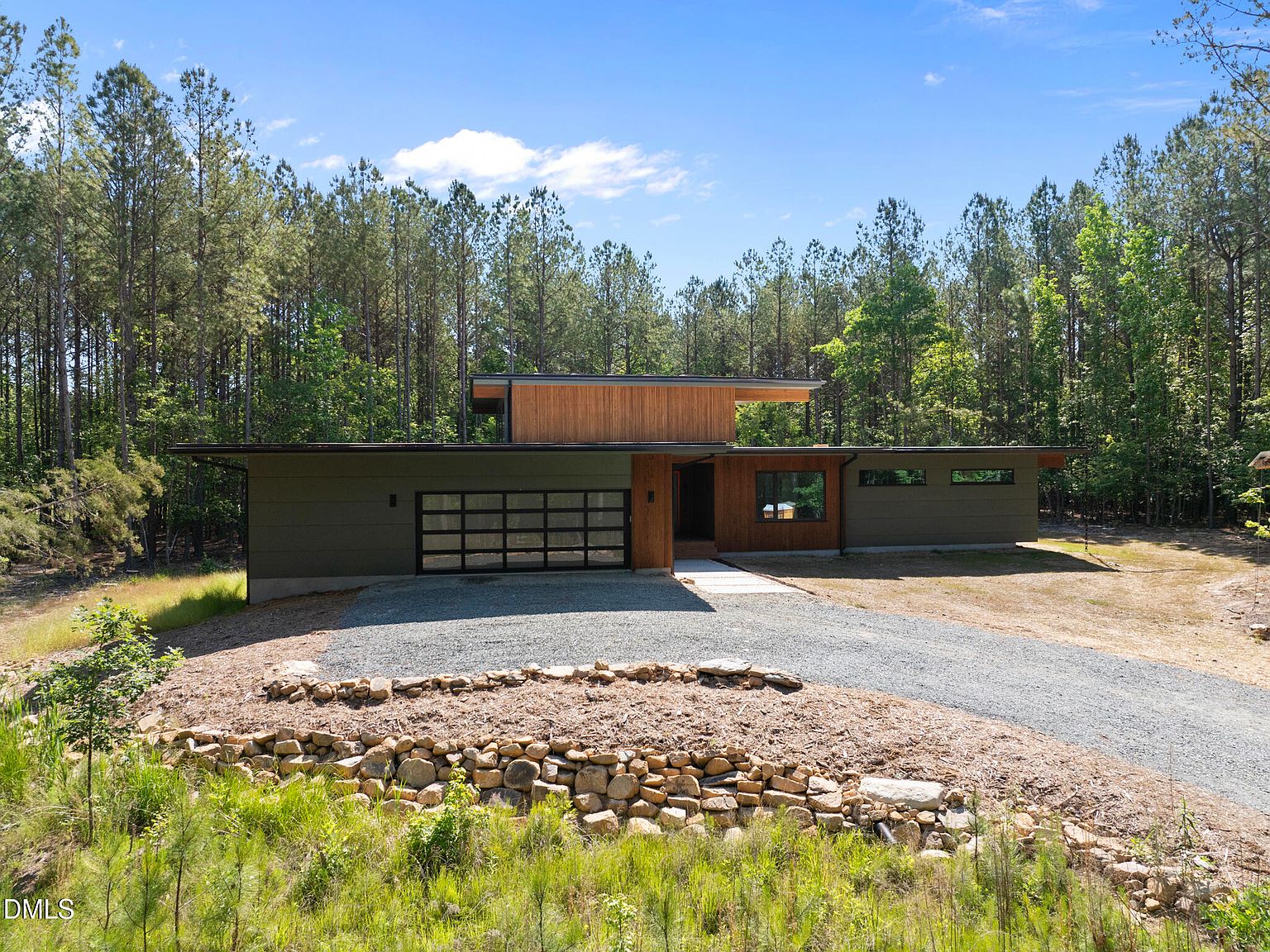 Daytime front elevation of 4637 Array Dr, a low-profile modernist ranch in cedar and dark green siding, set back from a gravel drive with a circular stone dry creek in the foreground