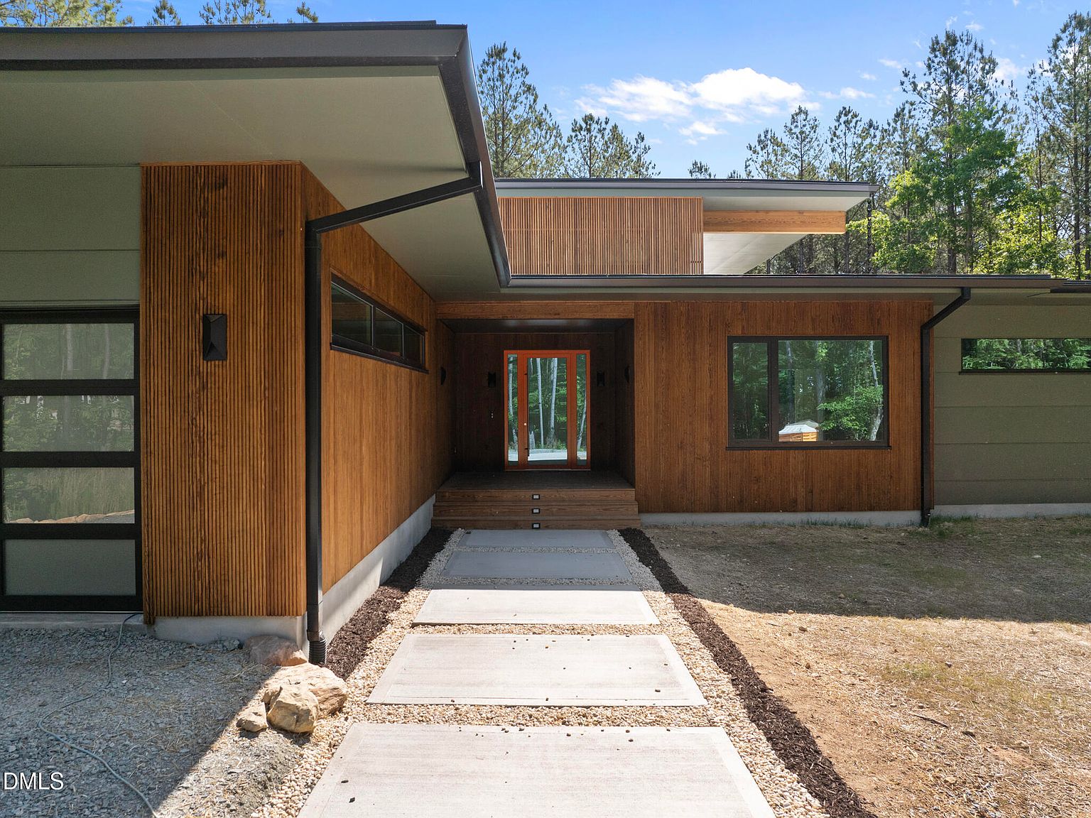 Close view of the cedar-clad entry: vertical wood siding, oversized glass front door, and a concrete-paver path framed by pea gravel and clipped grasses
