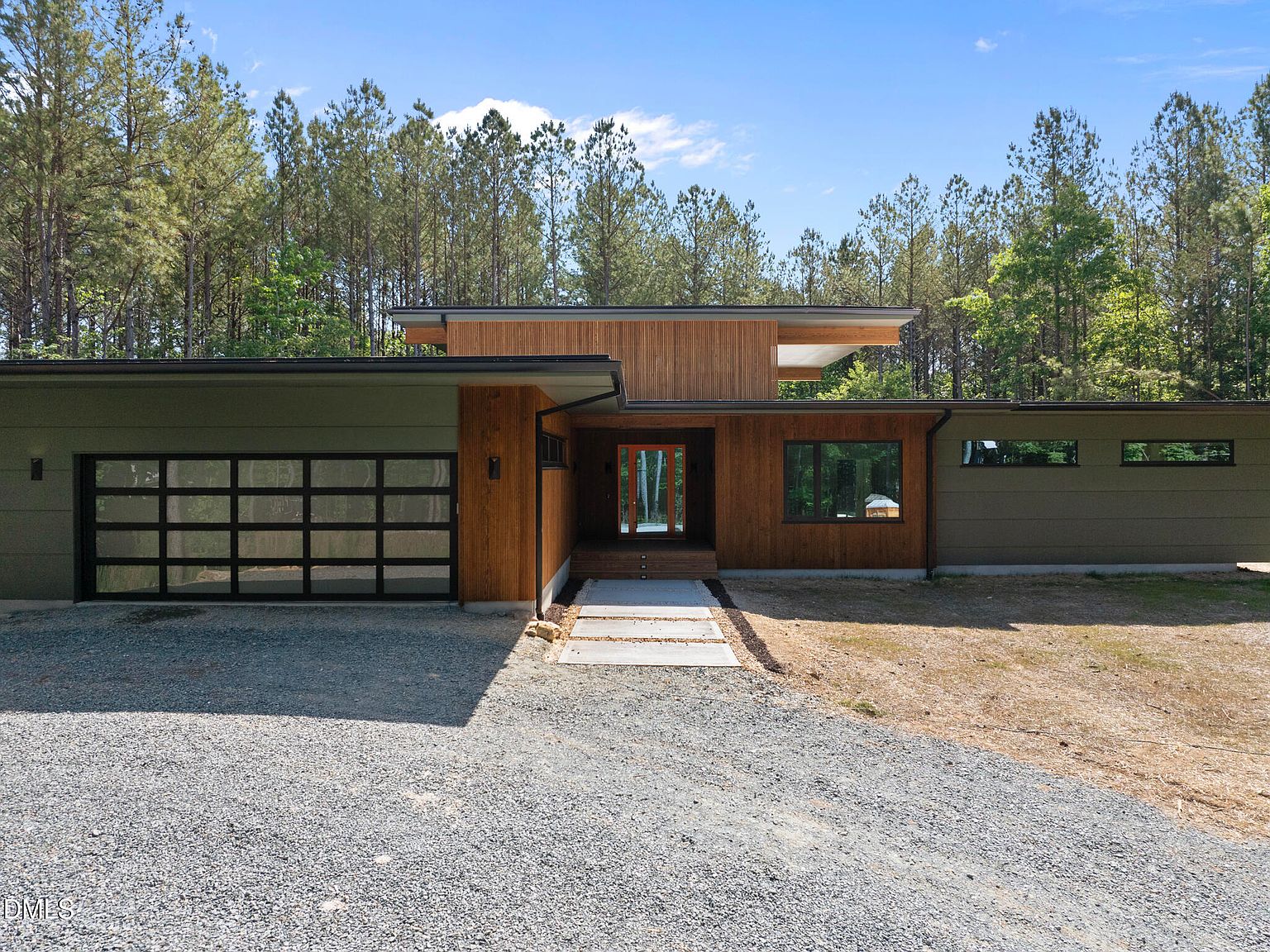 Wider three-quarter daytime view of the residence showing the deep eaves of the shed roof, glass-paneled garage door, and the cleared lot in the surrounding pine forest